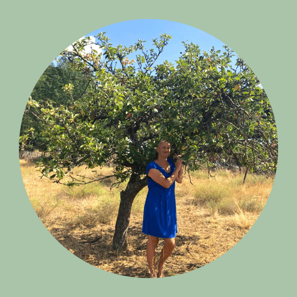 Photo of Kes Otter Lieffe, author of utopian trans speculative fiction novels and Queer Ecology writer. Kes stands in a blue dress beneath an apple tree in a grassy field